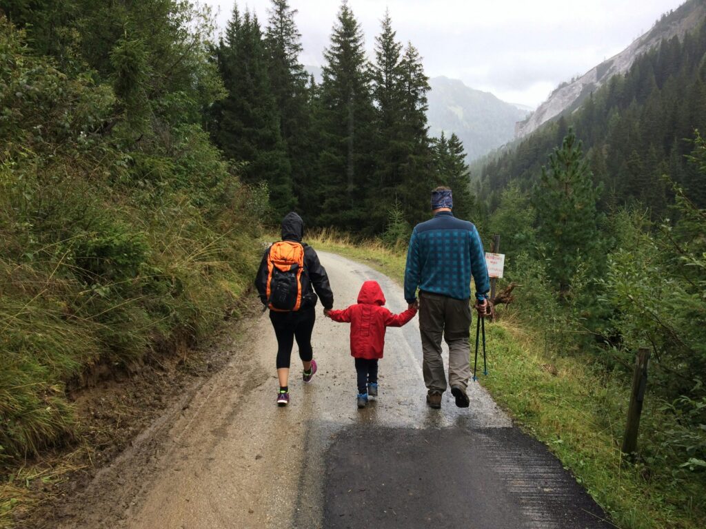 Family walking on a forest trail in the mountains, with parents holding hands with their child, all wearing outdoor gear.